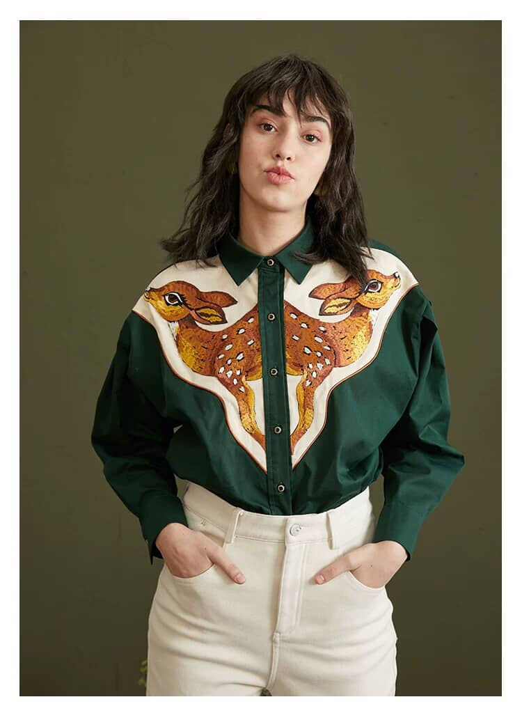 A woman wearing a dark green long-sleeve button-up shirt with a large embroidered deer design across the chest, paired with cream-colored pants, set against a solid dark olive green background.