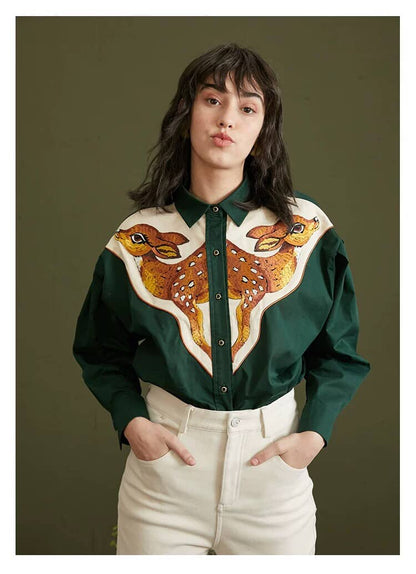A woman wearing a dark green long-sleeve button-up shirt with a large embroidered deer design across the chest, paired with cream-colored pants, set against a solid dark olive green background.